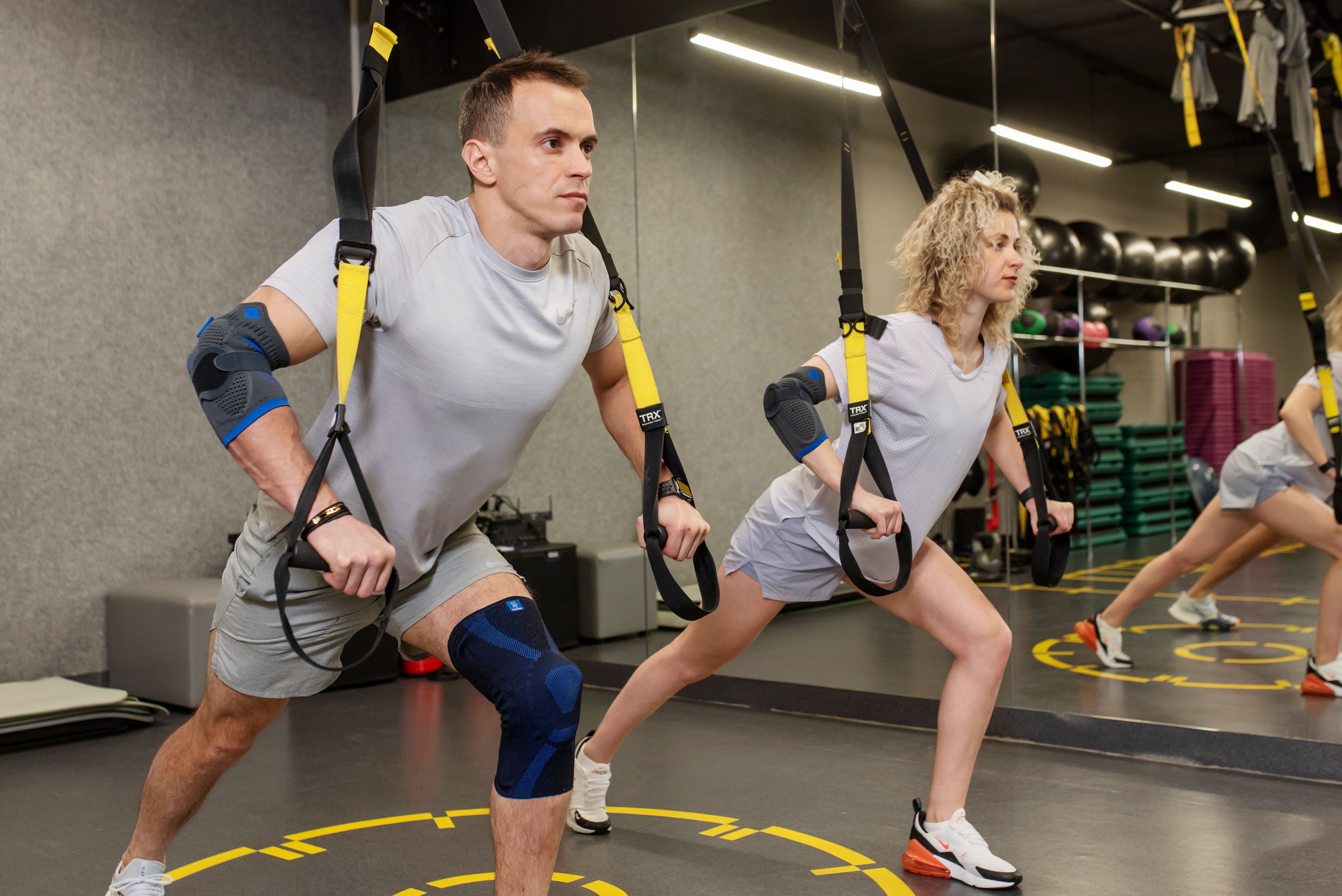 Чоловік та жінка тренуються з петлями TRX у ліктьових бандажах для захисту суглобів під час занять спортом.