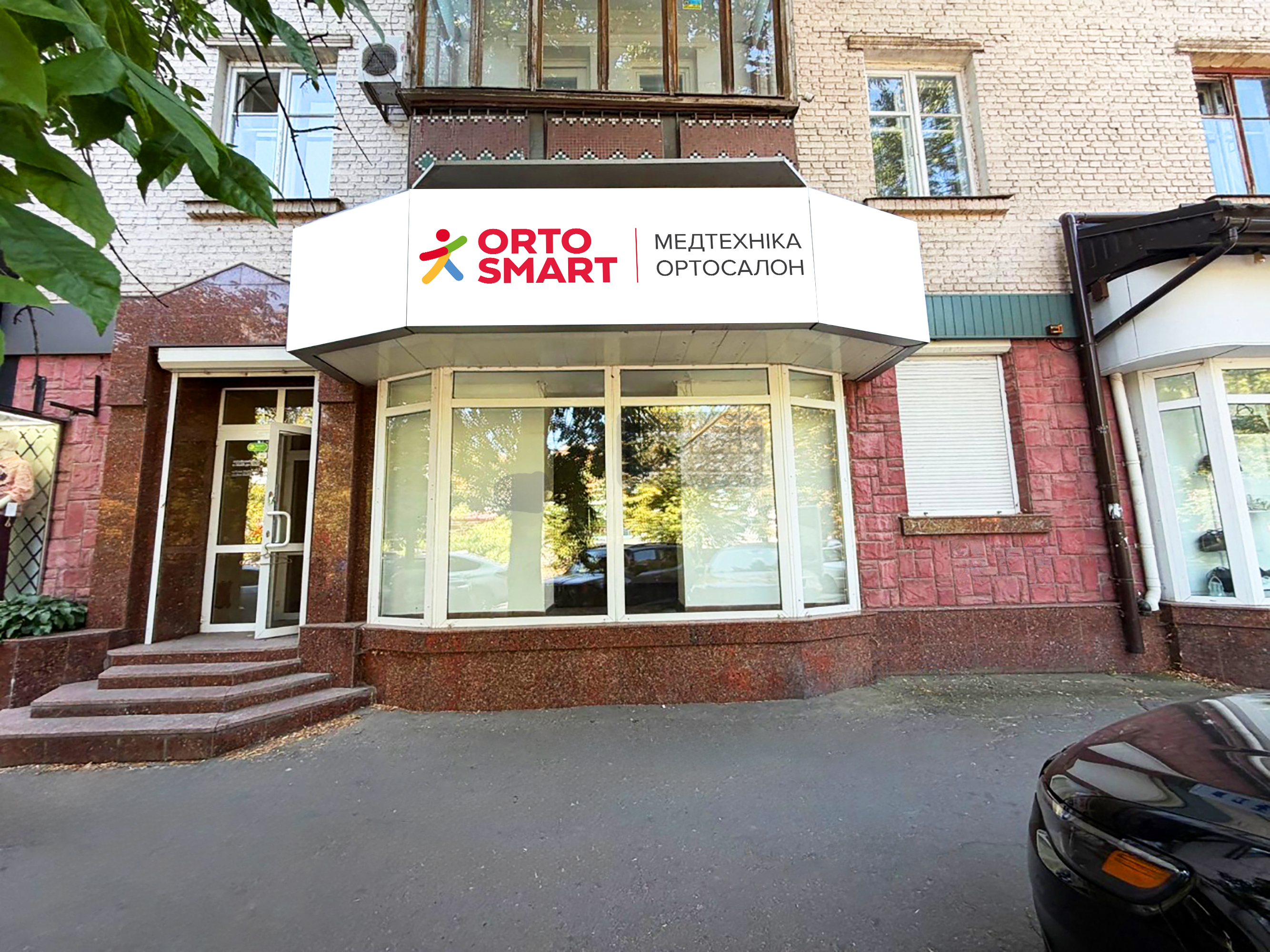 Широкоугольное уличное фото улучшенного и обновленного магазина ORTOSMART Медтехника Ортосалон в Черкассах. Магазин теперь открыт, с УБРАННОЙ роллетой. Большая угловая вывеска ORTOSMART (как на предыдущем фото) четкая и чистая. Стеклянные витрины чистые, с видимыми товарами и манекенами. Плитка, окна и ступени отремонтированы. Здание с чистой кирпичной кладкой и новыми оконными рамами. Зеленые деревья на переднем плане и черный автомобиль справа. Чистый тротуар.